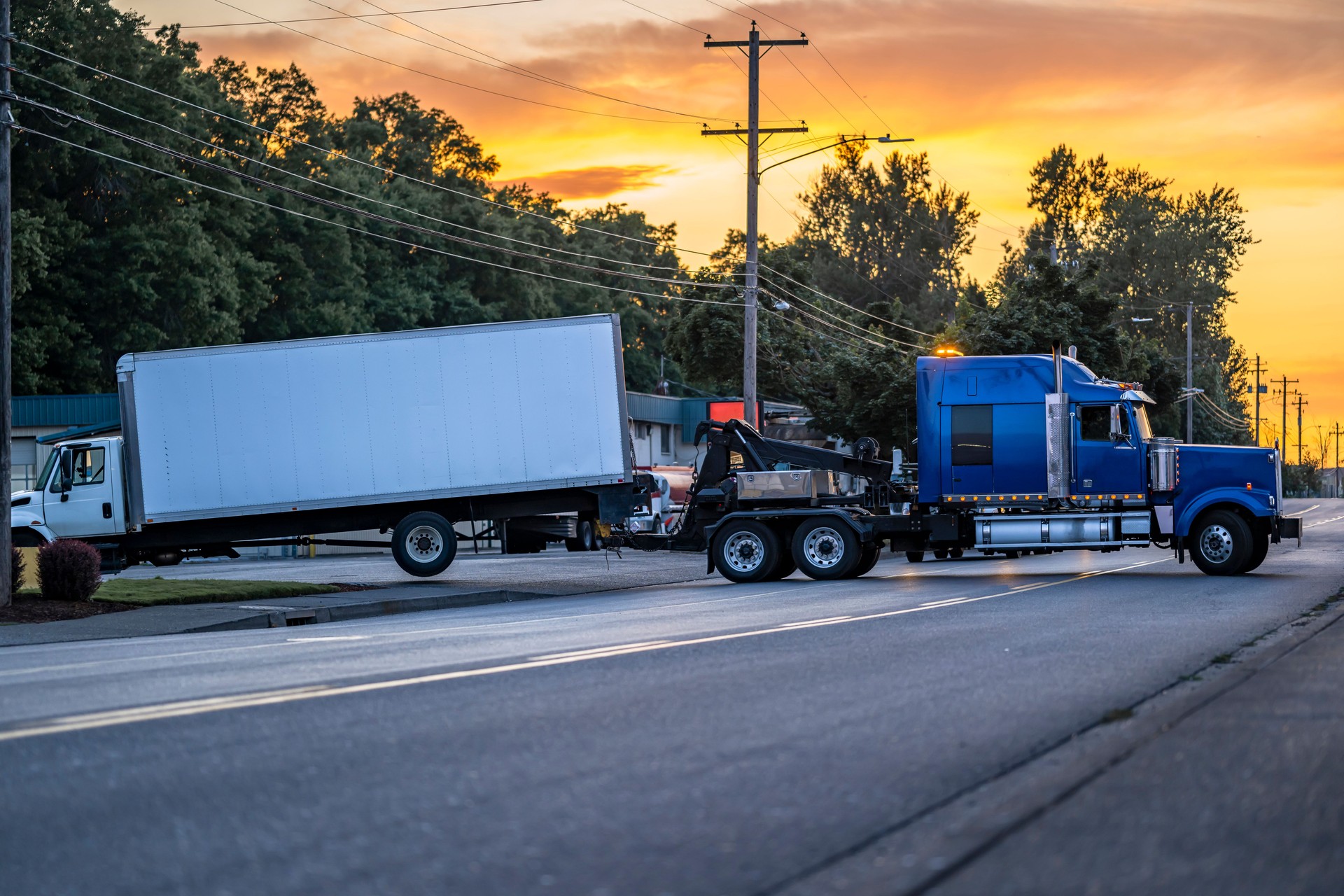 Heavy duty blue big rig tow semi truck towing broken semi truck with box trailer turning on the local road at sunset time